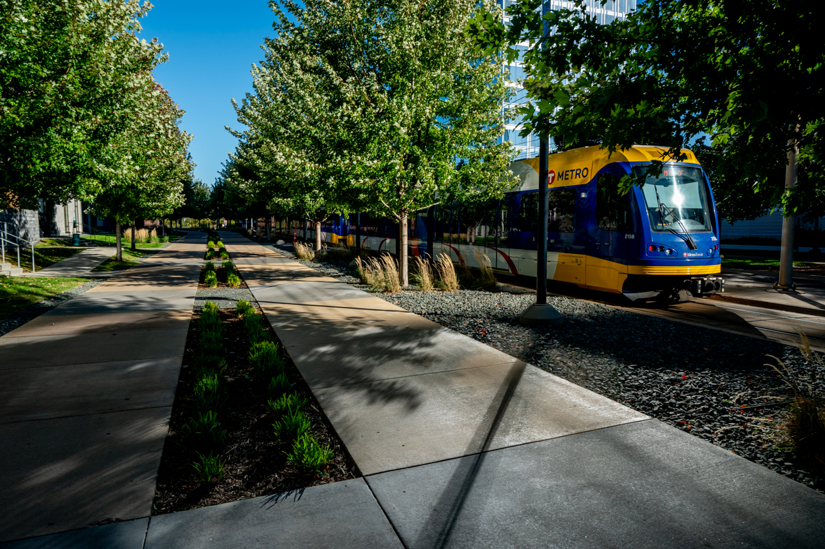 A Blue Line train travels through Bloomington Central Station Park, shaded by trees, with walking paths in the foreground and glassy buildings in the background.